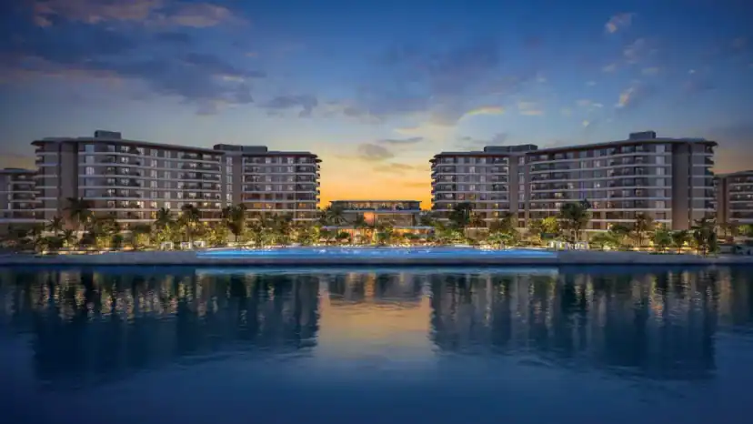 Two wide residential buildings with lit balconies face a waterfront at dusk, with a pool and palm trees between them and reflections on the water.