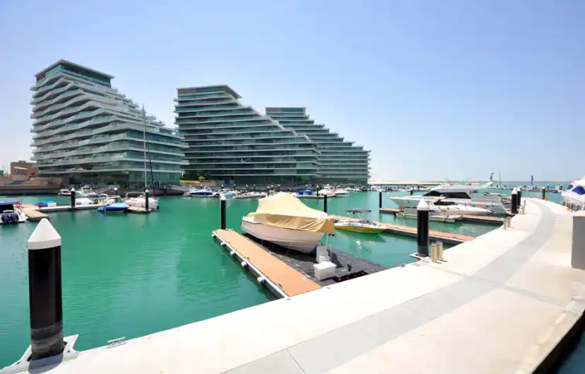 Marina with a covered boat at a wooden floating dock and yachts in turquoise water, with three terraced glass buildings in the background.