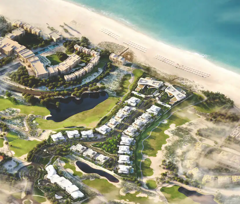 Aerial view of a beachfront development with low white buildings, villas among green fairways, two dark ponds and rows of loungers on a wide sandy beach.