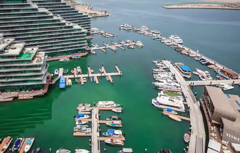 Aerial view of a marina with many small boats and yachts moored at floating docks beside a tall terraced glass building on green water.