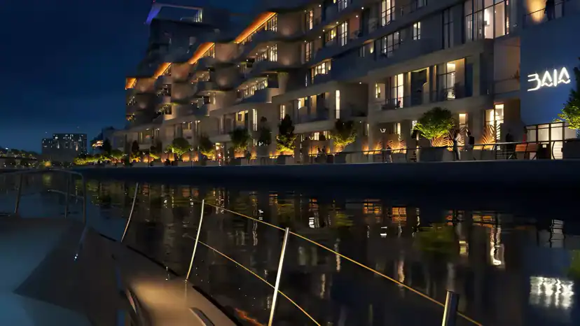 Night view from the water of a stepped waterfront apartment building with lit balconies, warm lights and planted trees along the promenade, and a sign on the right.