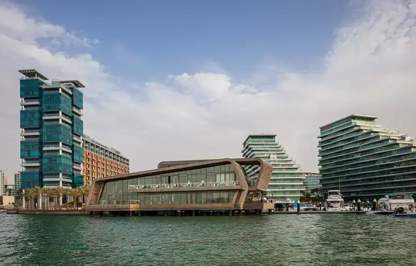 Waterfront view of a low curved building with large windows and a wooden-toned exterior, beside tall glass towers and terraced buildings under a cloudy sky.
