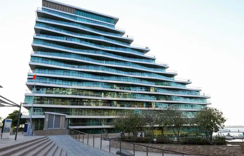 Tall residential building with stepped, sloping glass balconies seen from a paved waterfront walkway with stairs, a flag and a few trees.