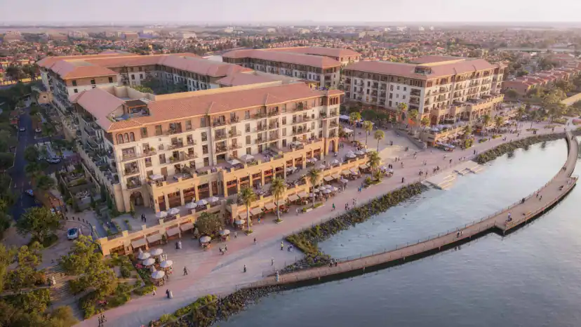 Aerial rendering of a terracotta-roofed apartment complex beside a curved waterfront with a wooden boardwalk, palm trees, outdoor seating, and surrounding low-rise houses.