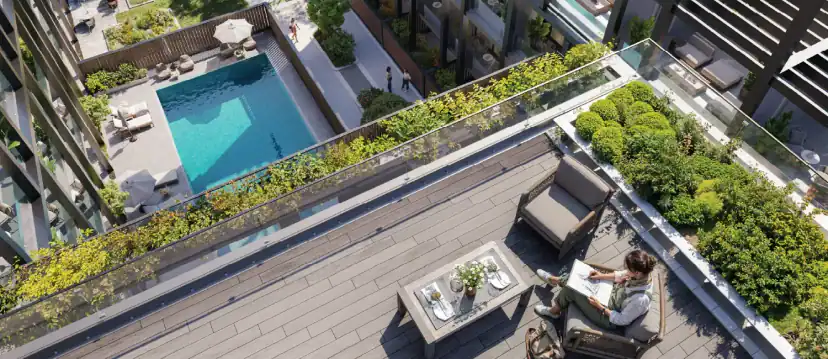Aerial view of a rooftop wooden deck with a chair, table and planters, and a person reading, above a swimming pool in a courtyard.