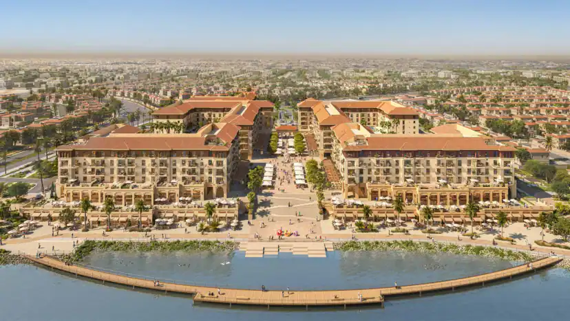 Aerial rendering of two large courtyard buildings with terracotta roofs facing a waterfront, with a central pedestrian plaza, a wooden boardwalk, and houses beyond.